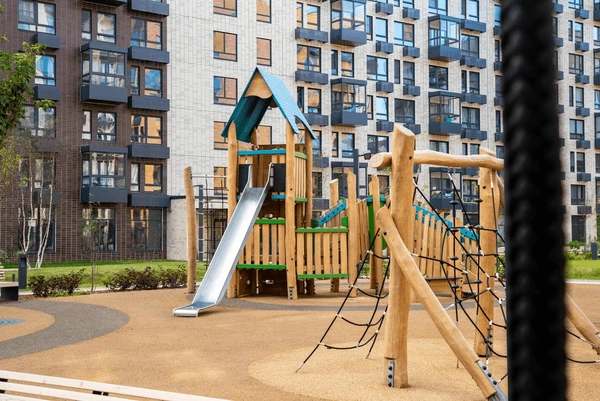 Wooden playground with a slide in a residential courtyard near modern apartment buildings on a sunny day.