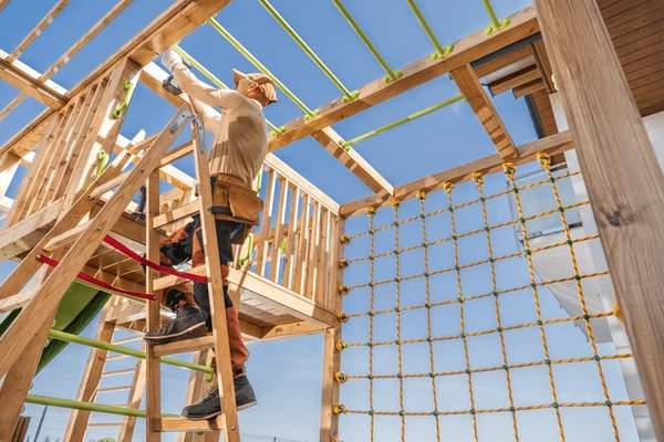 Worker installing a timber playground structure with a rope climbing net and overhead beams