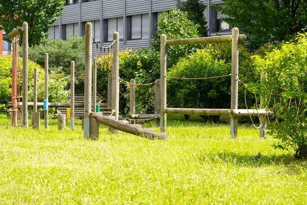 Wooden playground obstacle equipment on a grassy lawn, surrounded by green shrubs and trees near a building.