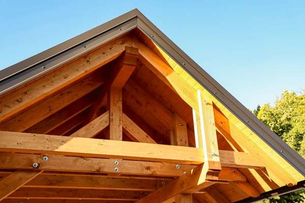 Close-up of a wooden roof structure with exposed beams and triangular framing under a sloped roof, lit by sunlight against a clear blue sky.