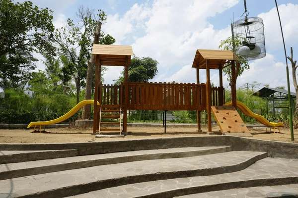 Wooden outdoor playground with yellow slides, a climbing ramp, and steps, set on a sandy area beside tiered stone seating under a partly cloudy sky.