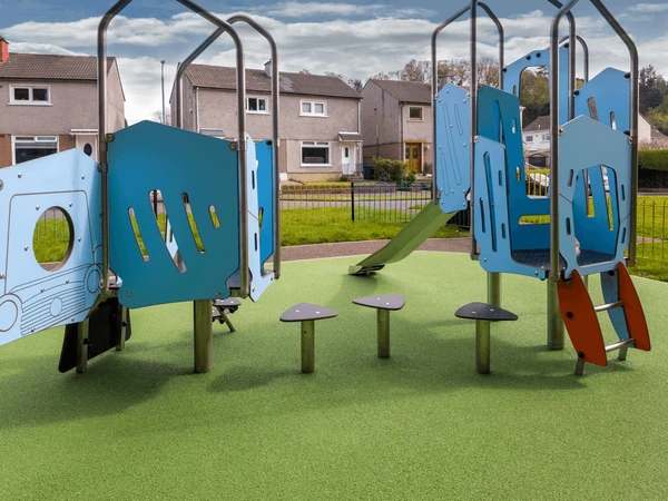 Blue playground structures with ramps, stepping platforms, and railings sit on a green rubber play surface, with houses and a grassy area in the background.